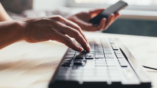 Image showing an identifiable person's hands typing on a keyboard and holding a phone with the other hand.