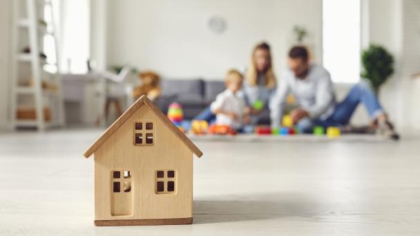 Parents and young children at home in background, blurred. In the foreground a small toy wooden house. 